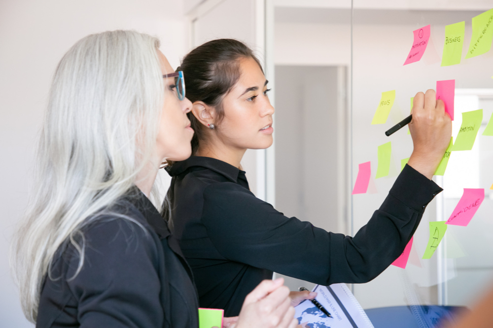 confident-latin-businesswoman-writing-stickers-sharing-ideas-project-focused-grey-haired-female-manager-reading-notes-glass-wall